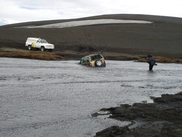 Green SUV stuck in a river with a person wading nearby and a white truck on the bank