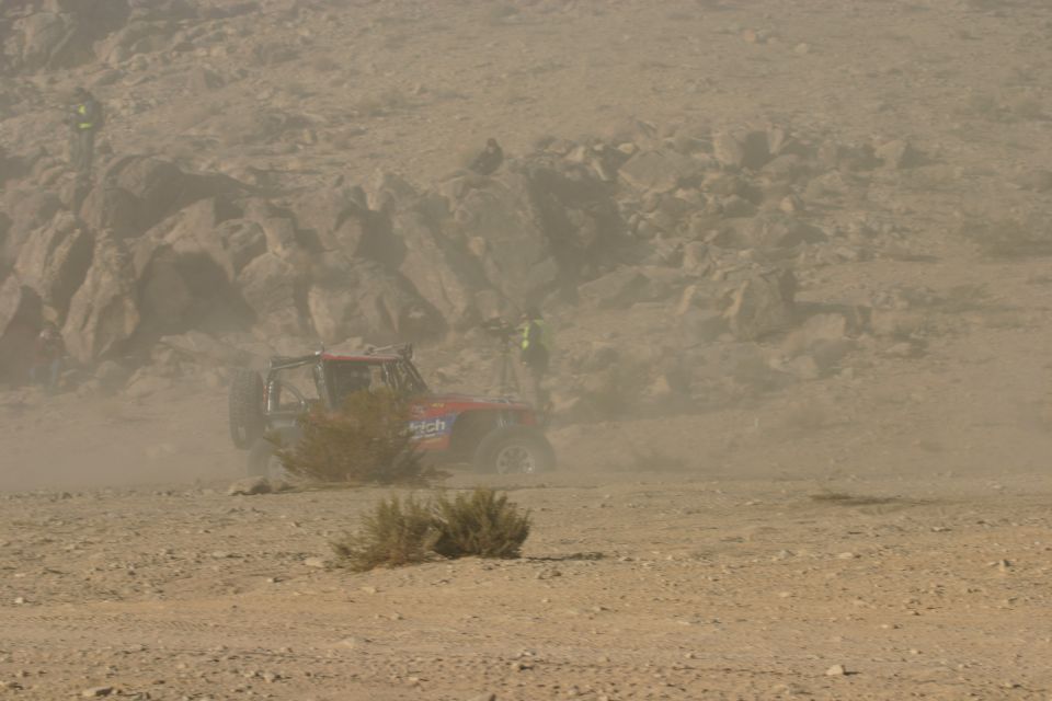 Off-road buggy kicking up dust on a desert course. Some people stand on rocks in the background