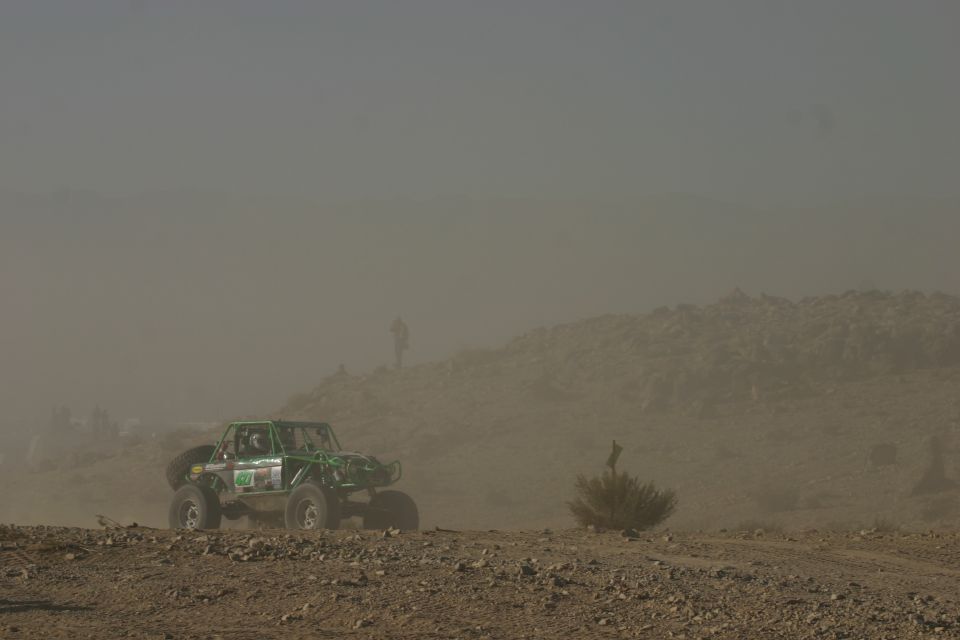 Green off-road buggy kicking up dust on a rocky desert terrain. A person stands on a distant hill