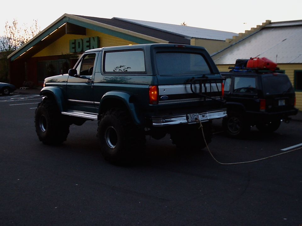 Lifted teal Ford Bronco on big tires towing a dark Jeep Cherokee in a parking lot at dusk.
