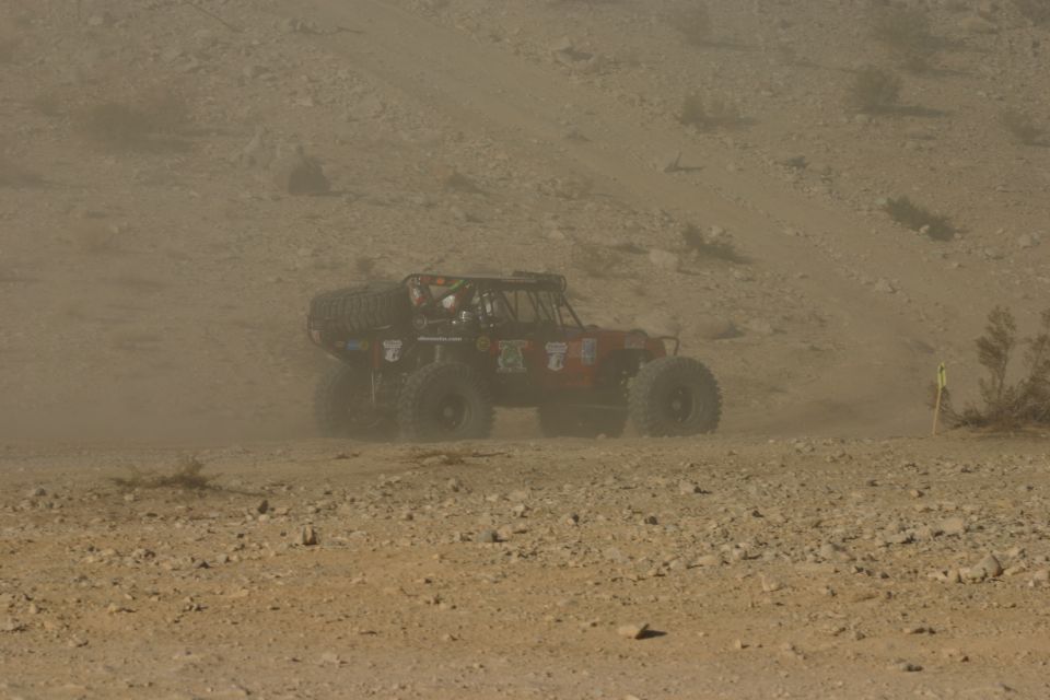 Buggy with large tires driving on a dusty, rocky hill. It has a spare tire on the back and several decals on its side