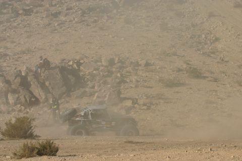 1992 Jeep Wrangler - A custom off-road buggy navigates a dusty desert terrain during a competition, with spectators observing from a nearby rocky area.