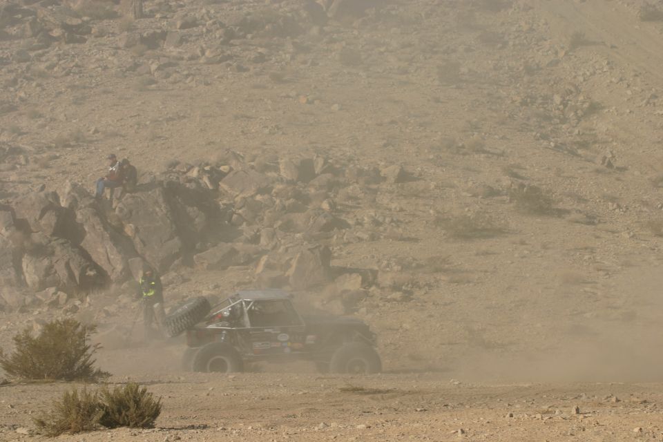 Off-road buggy kicking up dust in a desert race. Two people watch from a rocky outcrop