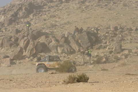 1992 Jeep Wrangler - A yellow off-road buggy kicks up dust as it navigates a desert landscape, with people watching from a rocky outcrop.