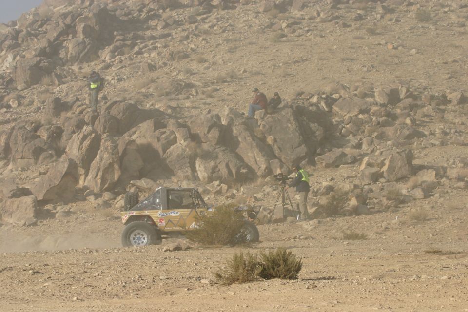 Yellow off-road buggy with roll cage, driving on a dusty desert terrain with spectators on a rocky hill