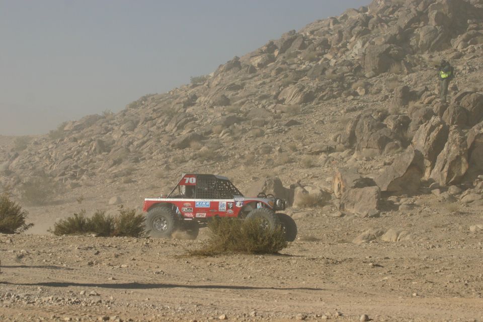 Red off-road buggy with number 70 on roof, driving on dusty desert terrain with rocky hills in background