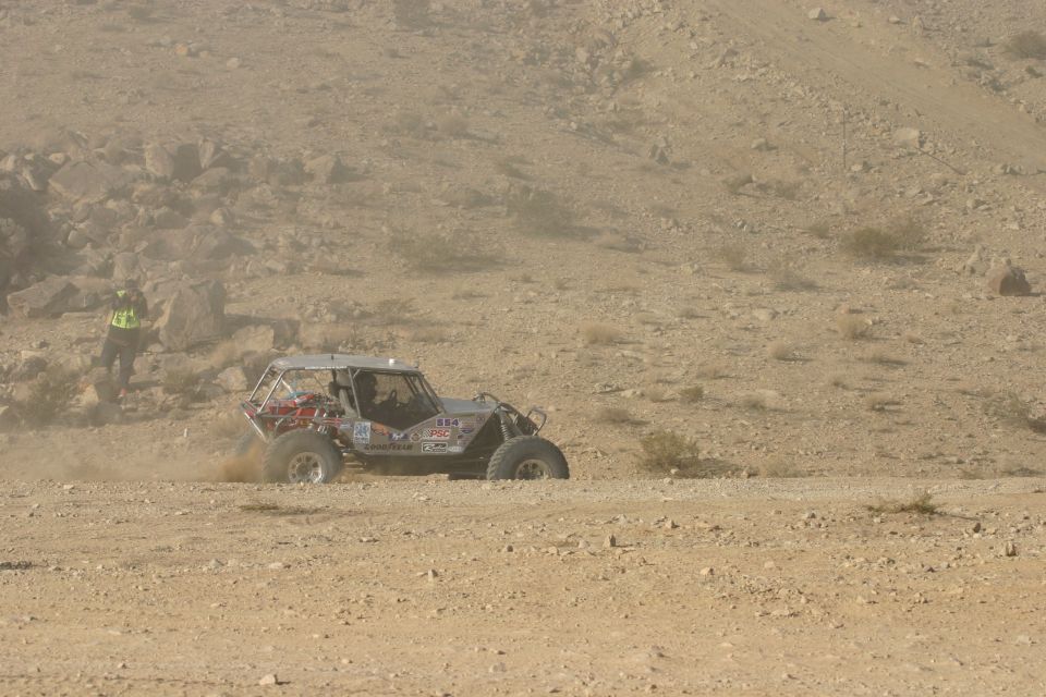 Off-road buggy with roll cage kicking up dust on a dirt track in a desert landscape. A person in a high-visibility vest stands to the left