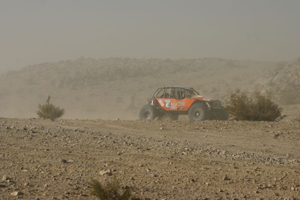 Orange off-road buggy with roll cage driving on a dusty desert trail. Some bushes and rocky hills in background