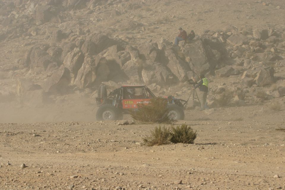 Off-road buggy kicking up dust on a desert course, with spectators on a rocky hillside