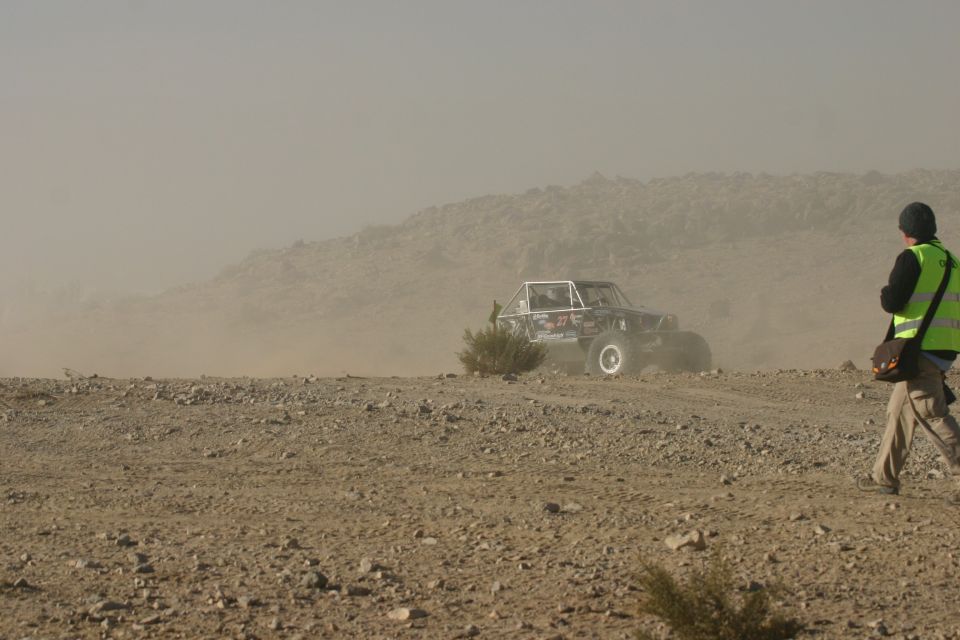 Off-road buggy with roll cage driving through dust, person in high-visibility vest walks nearby