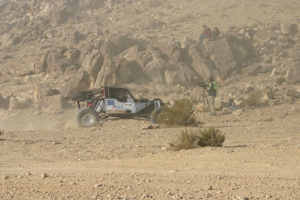 Off-road buggy with number 366 on side, kicking up dust on a desert race course. A cameraman stands nearby