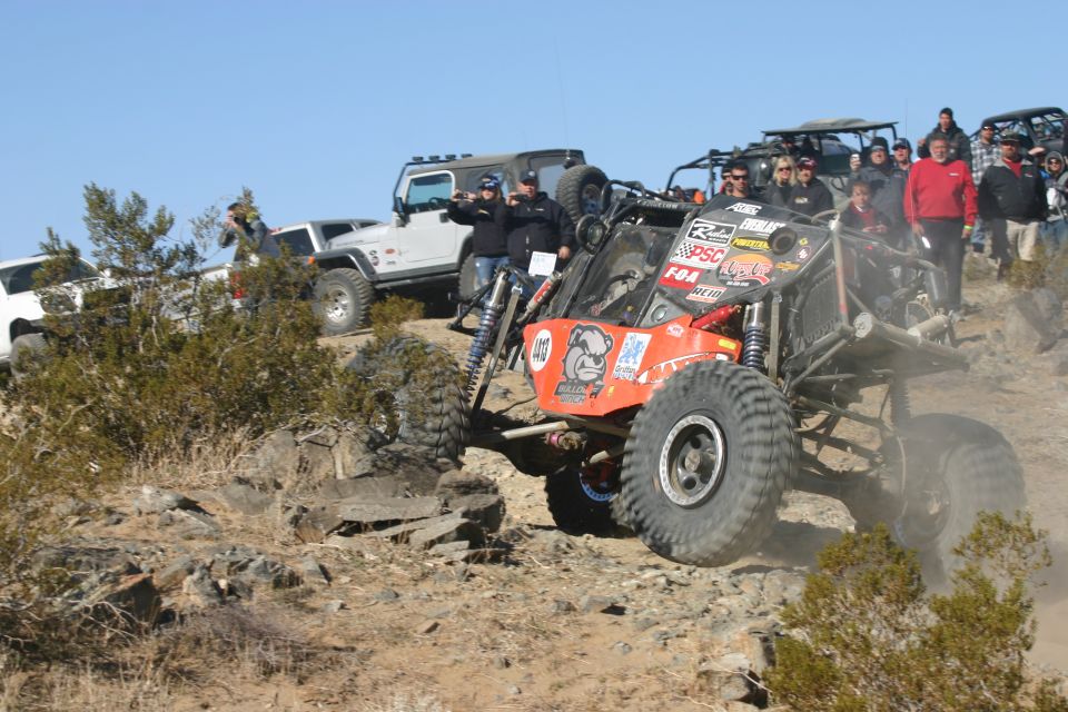 Orange off-road buggy with large tires climbing a rocky incline, other Jeeps and spectators in background