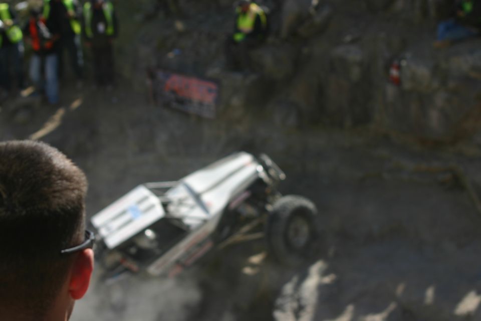 Off-road buggy with white body and roll cage climbing a steep, rocky incline. Spectators are visible on the left