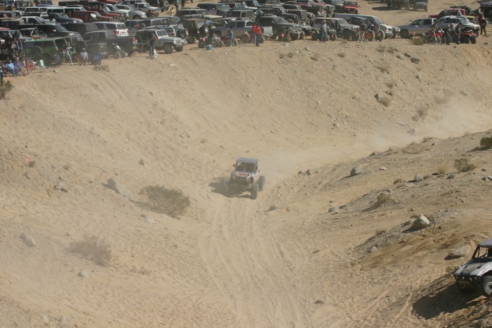 Off-road buggy driving down a dusty sand dune, with many parked vehicles on the ridge above