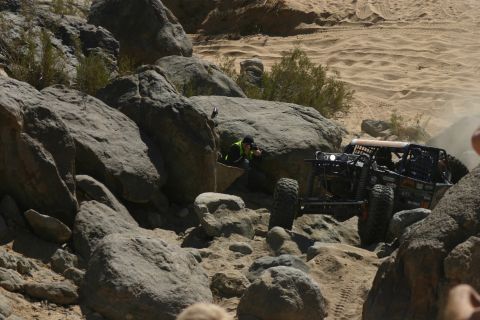 1992 Jeep Wrangler - off-road rock crawler buggy with large tires negotiates a boulder field in a sandy desert while a person in safety vest spots.