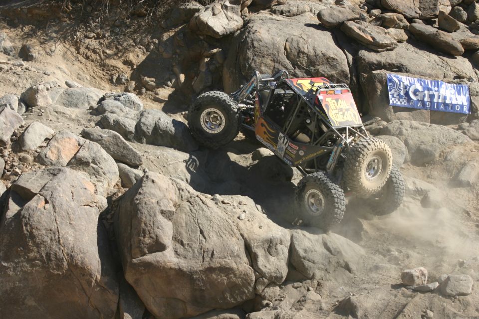 Off-road buggy with "TRAIL GEAR" sticker on a rocky incline, kicking up dust. Banner for "CITIZEN RADIATOR" visible