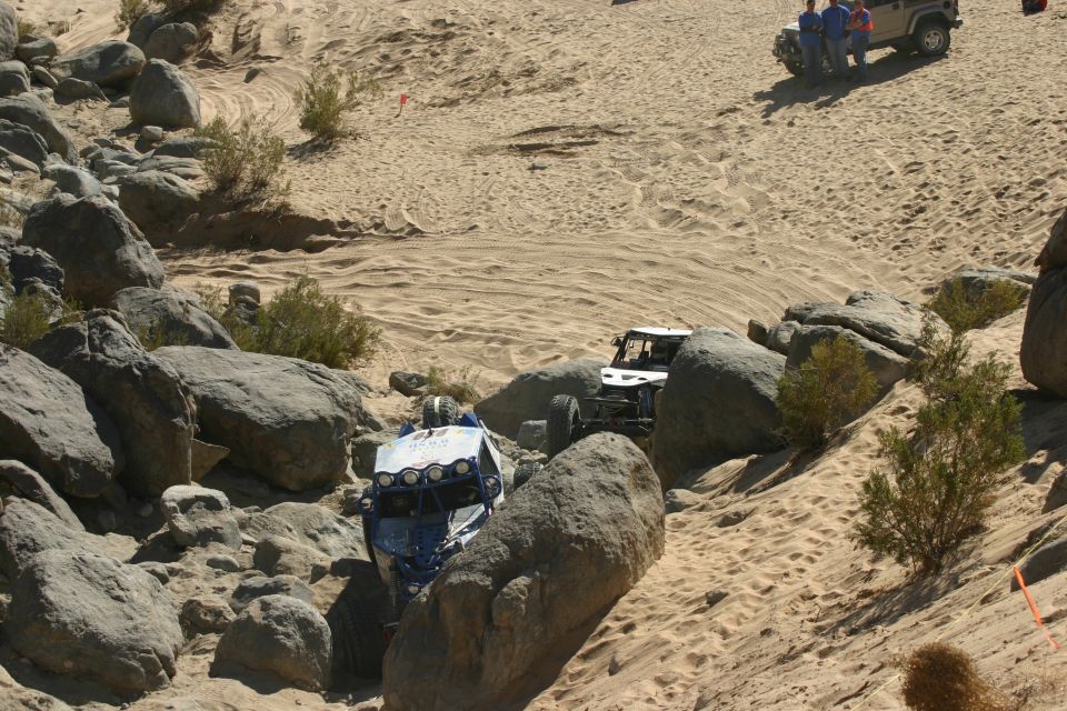 Two off-road vehicles navigate a rocky, sandy terrain with sparse desert vegetation