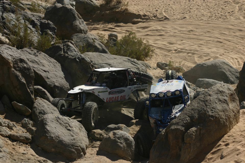 Two off-road buggies with sponsor decals driving through a rocky desert landscape. One buggy is white with Lucas Oil branding, the other is blue