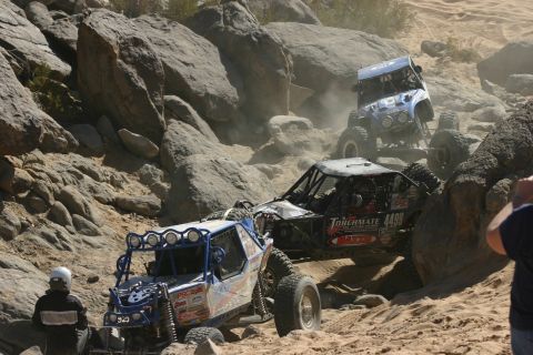 1992 Jeep Wrangler - Multiple custom off-road vehicles tackle a challenging rocky terrain, kicking up dust as they ascend.