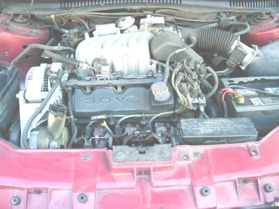 1997 Ford Taurus - Engine bay of a red Ford Taurus showing the Vulcan V6 with "3.0 V6" cast into the black valve cover, upper intake plenum, and a conical ope…
