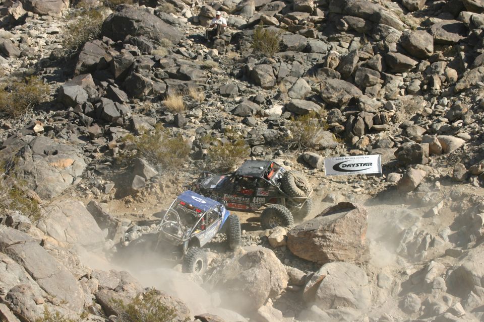 Two off-road buggies climbing a rocky hill, one blue, one black with spare tire