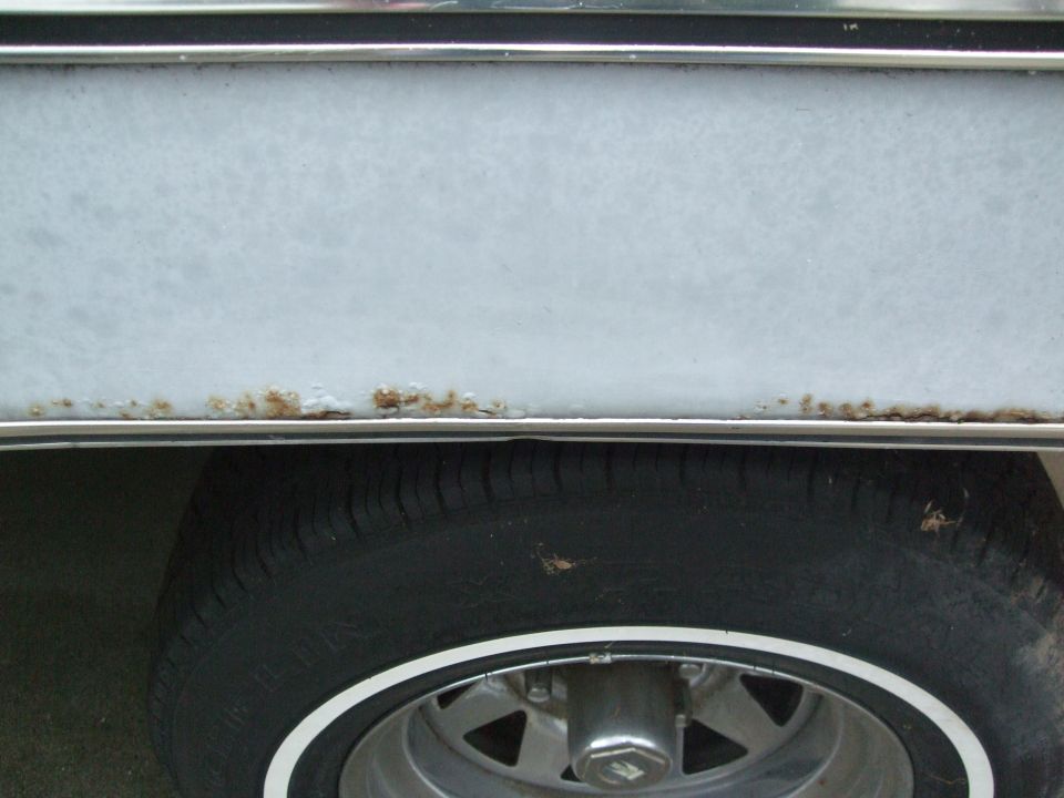 Light blue vehicle lower body panel with rust spots above a tire with a white stripe on the rim