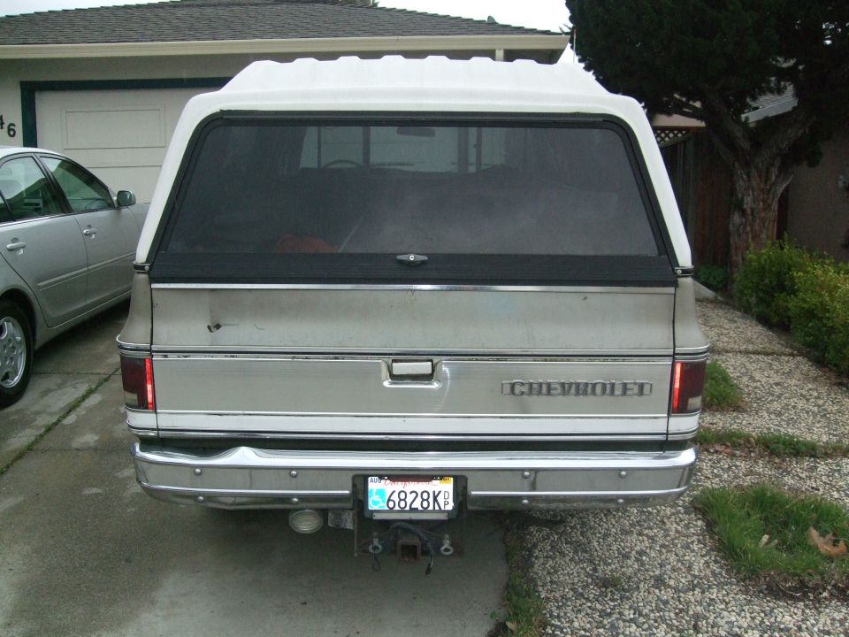 Rear view of a Chevrolet pickup truck with a white camper shell. Another car is parked to the left