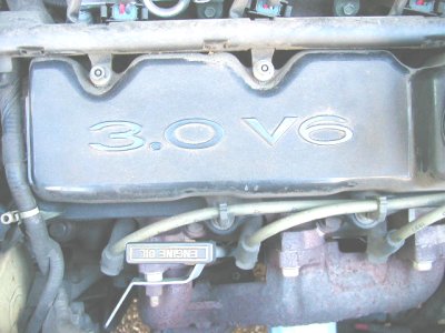 1997 Ford Taurus - Overhead close-up of a dusty engine bay showing a valve cover cast with '3.0 V6' lettering, with the intake manifold tube above it, ignitio…