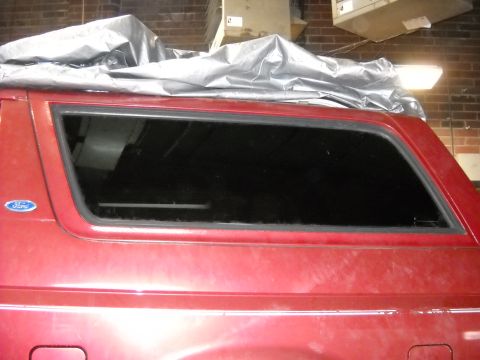 1993 Ford C-150 (Centurion Conversions) - The rear side window of a red SUV is visible, with a tarp covering the roof and a brick wall in the background.