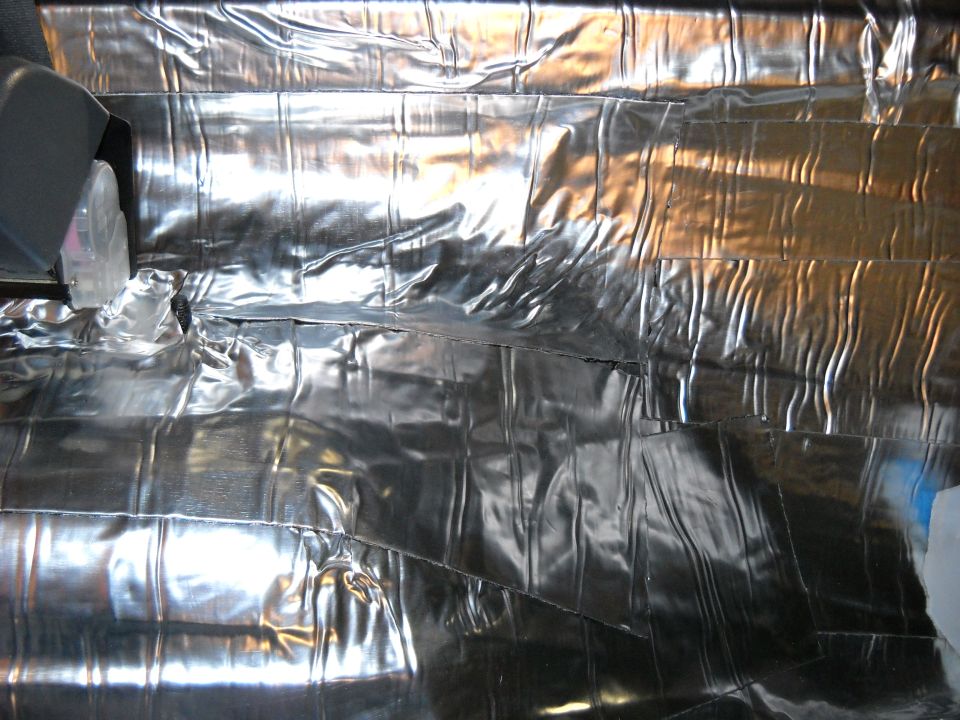 Interior of a vehicle with silver insulation covering the floor and walls. A grey plastic seat base is visible on the left