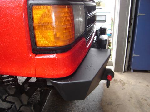 1989 Ford Bronco - The front corner of a red vehicle is visible, an amber turn signal and a black aftermarket bumper with a red tow hook.