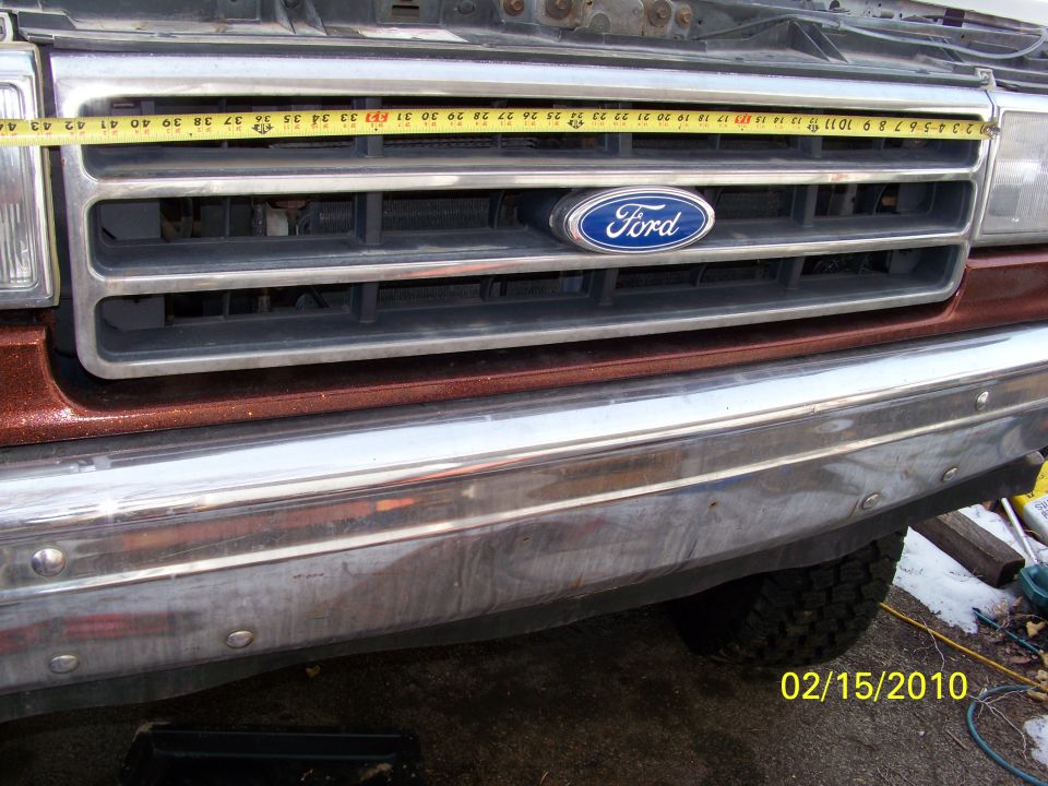 Close-up of a Ford truck grille with a tape measure across it. The grille has horizontal bars and a blue Ford oval emblem