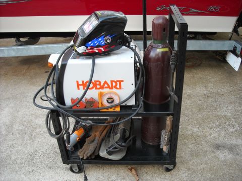 1979 Ford F-150 - A cart holds welding equipment including a Hobart welder, a helmet, and a gas cylinder, with a boat on a trailer in the background.