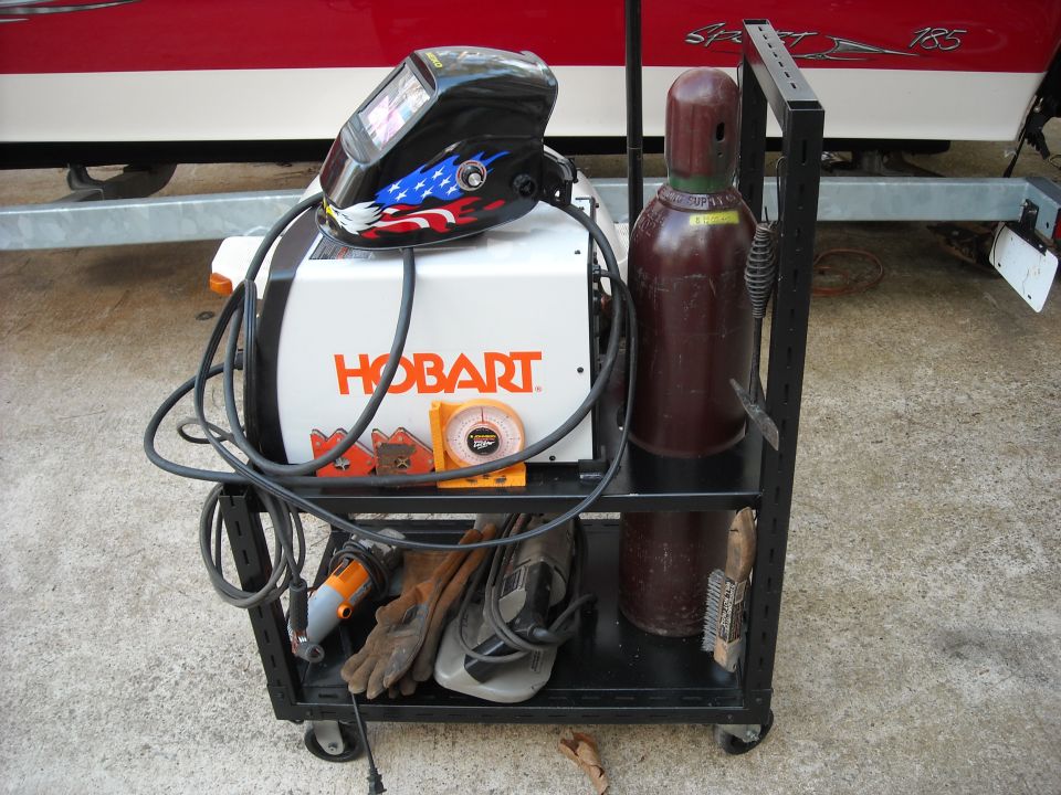 Hobart welder, welding helmet with American flag design, gas cylinder, gloves, and brushes on a cart