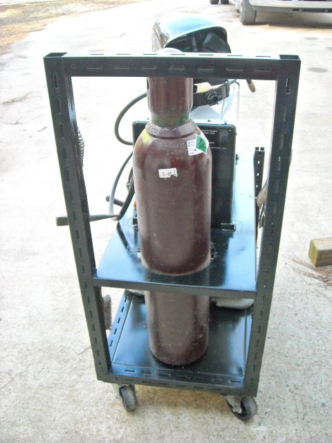 1979 Ford F-150 - A welding cart holds two gas cylinders and a welding helmet, with a portion of a vehicle visible in the background.