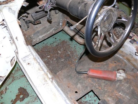 1976 Chevrolet Corvette - Stripped car interior showing the bare, rusty floor pan with teal-green original paint remnants, exposed brake/accelerator pedals, steering…