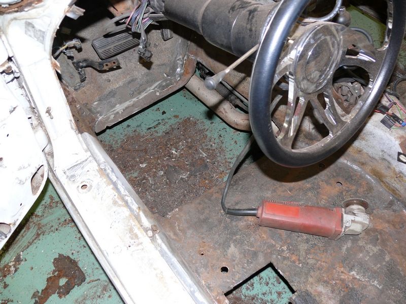 Bare rusty car floor pan with pedals, steering wheel and a red angle grinder