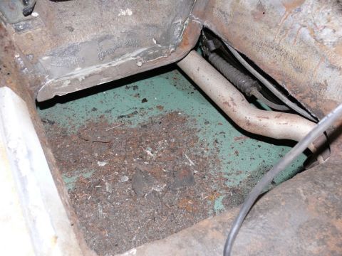 1976 Chevrolet Corvette - Interior floor pan viewed from above with green paint or coating partially covered by dirt, rust flakes and debris. A beige-painted metal t…