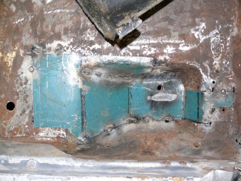 1976 Chevrolet Corvette - Underside/floor pan area of a vehicle showing bare and rusted sheet metal with fresh weld beads and a teal-painted reinforcement panel. Mul…