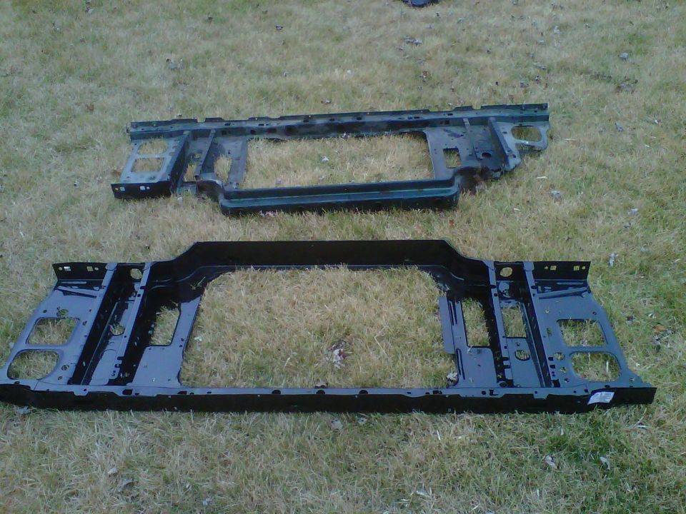 Two automotive radiator core support frames on grass, weathered gray above glossy black below