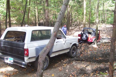 1989 Ford Bronco - A white Ford Bronco is stuck in the mud and being towed by a red Jeep in a forest setting.