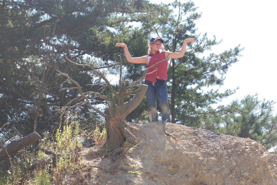Child in red vest and jeans stands on a dirt mound, arms outstretched, with trees in the background