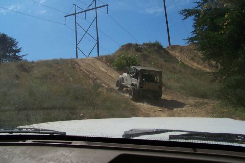 1989 Ford Bronco - A tan soft-top Jeep Wrangler on lifted suspension and knobby tires climbs a dusty dirt hill at an off-road park, photographed through the w…