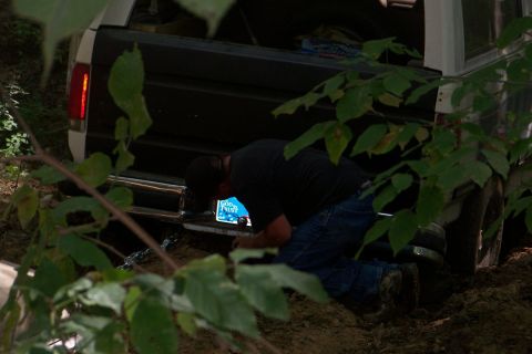 1989 Ford Bronco - Rear of a white full-size Ford SUV with the tailgate down, buried to the axle in dirt on a wooded trail while a man in a black t-shirt knee…