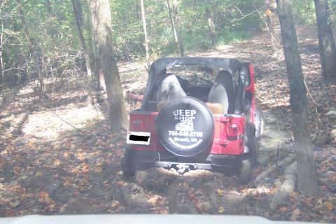 1989 Ford Bronco - Rear view of a red soft-top Jeep Wrangler working through a rutted, muddy trail in leafy woods, shot over the hood of a following vehicle.…