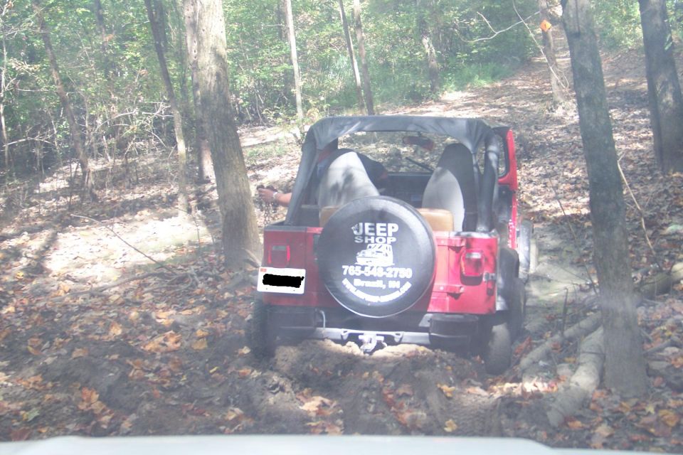 Red soft-top Jeep Wrangler on a muddy wooded trail with Jeep Shop tire cover