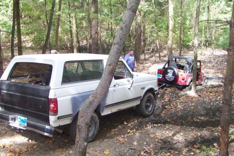 1989 Ford Bronco - White full-size Ford Bronco with a black primered tailgate and removable hardtop sits in a muddy wooded trail, viewed from the 3/4 rear dri…
