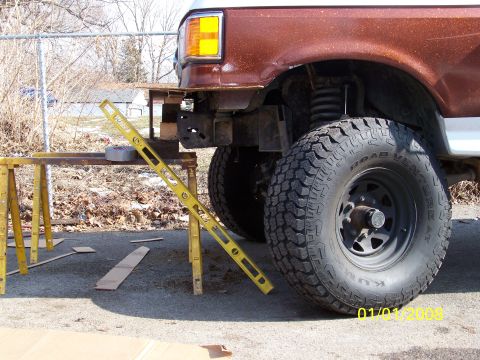 1989 Ford Bronco - Close-up of the front driver-side frame horns on a lifted brown Ford SUV with the bumper removed, showing the coil-spring front suspension…