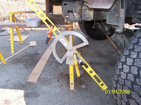 1989 Ford Bronco - A large protractor angle finder and a yellow level are propped against the front frame and skid plate of a lifted 4x4, used to measure cast…