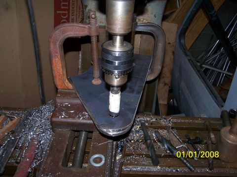 1989 Ford Bronco - A drill press fitted with a hole saw cutting a rounded steel bracket plate that is held to the table by two rusty C-clamps. The cast-iron t…